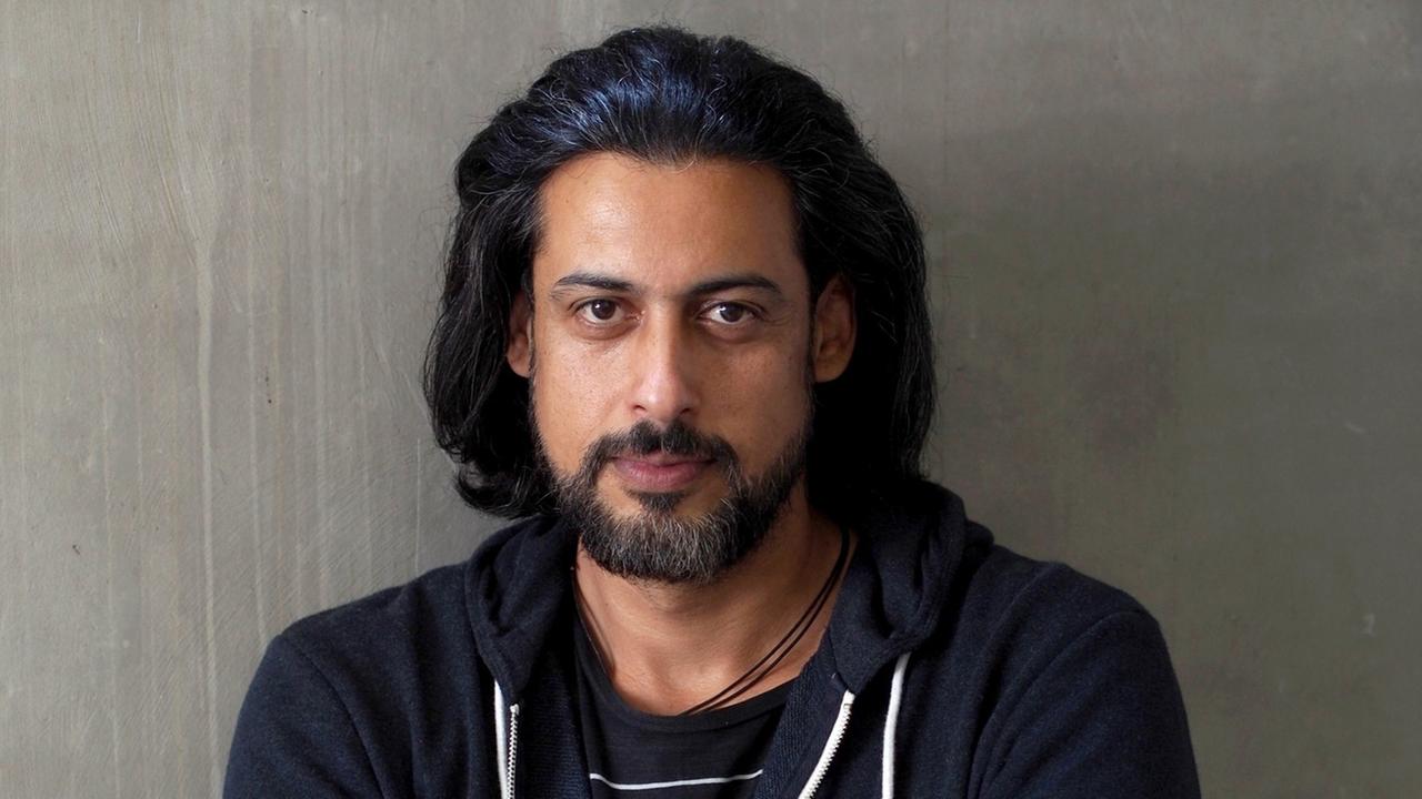 Abbas Khider steht vor einer grauen Wand. Er hält die Arme verschränkt.