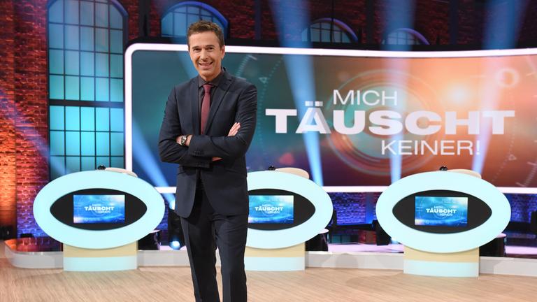 "Mich täuscht keiner! - Die Show der Sinne": Moderator Dirk Steffens in Ganzkörperansicht im Studio vor einem großen Monitor mit dem Sendungslogo und den Kandidatenpulten.