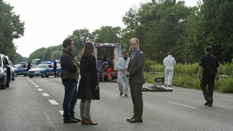 "Neben der Spur - Dein Wille geschehe": Ruiz (Juergen Maurer), Anna (Marie Leuenberger) und Joe (Ulrich Noethen) stehen auf einer abgesperrten Autobahn. Im Hintergrund stehen zwei Polizeiwägen und ein Krankenwagen sowie mehrere Beamte.