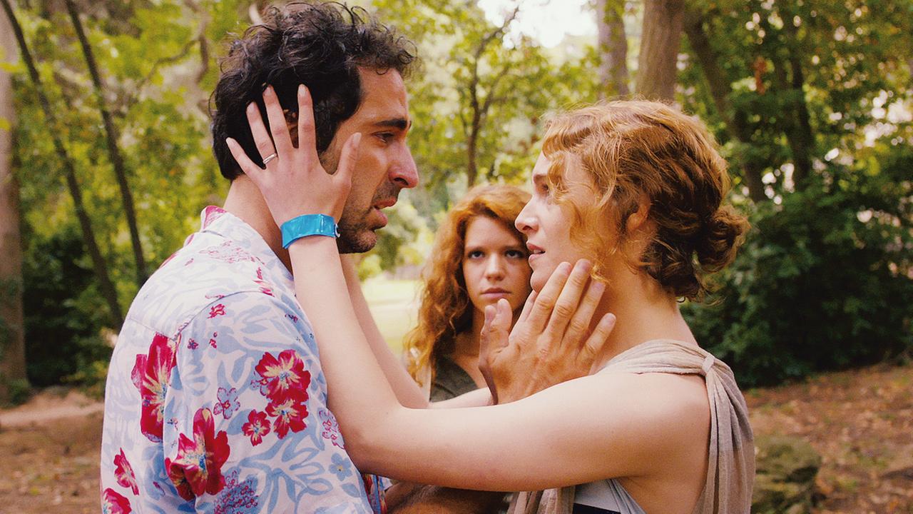 "Love Island": Grebo (Ermin Bravo) und Liliane (Ariane Labed) stehen voreinander in einem Wald. Sie halten mit ihren Händen jeweils das gesicht des Anderen fest. Im Hintergrund, zwischen den beiden, steht Flora (Ada Condeescu) und schaut die beiden an.