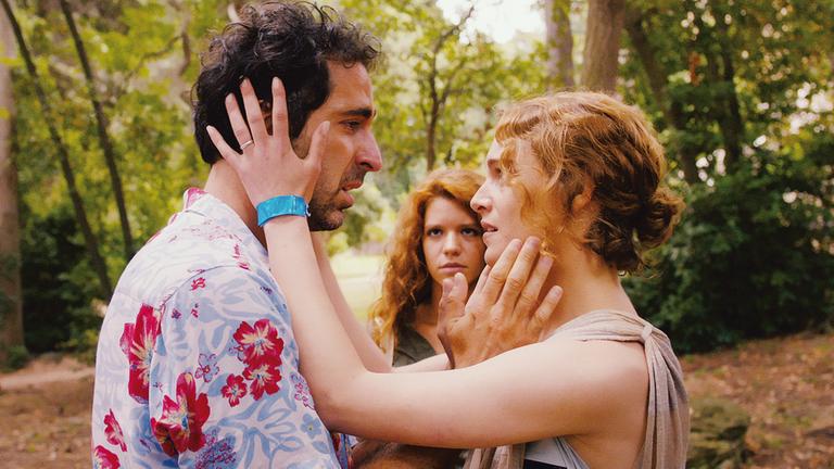 "Love Island": Grebo (Ermin Bravo) und Liliane (Ariane Labed) stehen voreinander in einem Wald. Sie halten mit ihren Händen jeweils das gesicht des Anderen fest. Im Hintergrund, zwischen den beiden, steht Flora (Ada Condeescu) und schaut die beiden an.