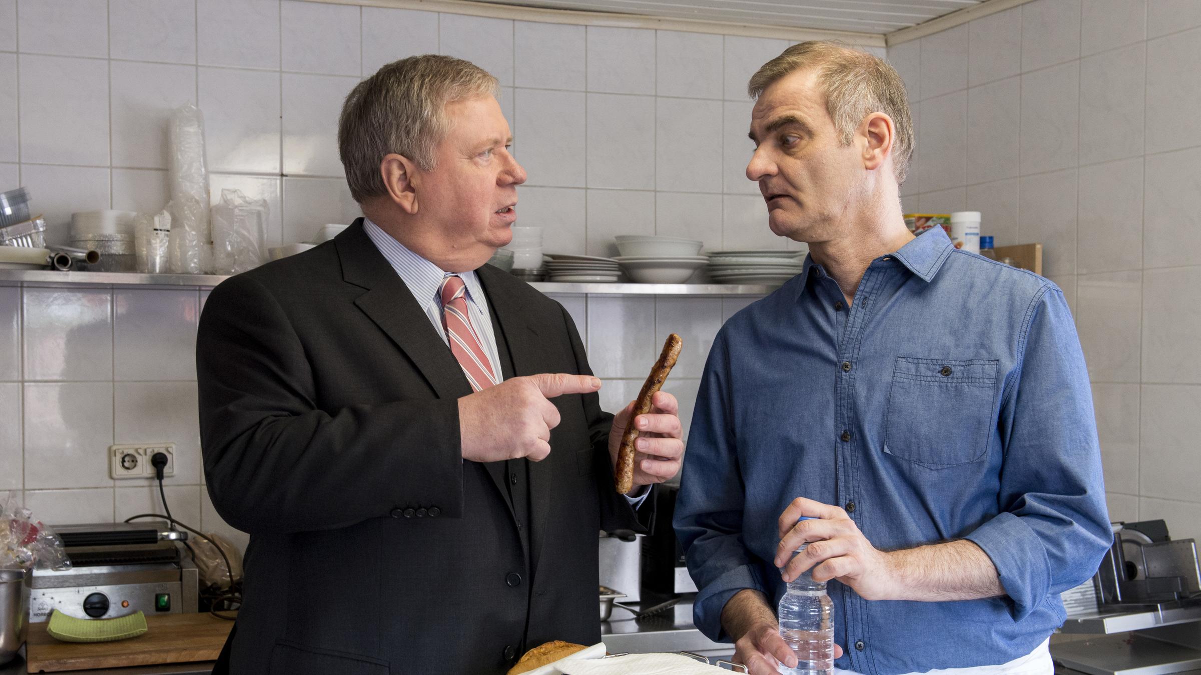 "Der Staatsanwalt – Tödliche Übernahme“: Im Imbiss erklärt Stefan Schiller (Heinrich Schafmeister) Staatsanwalt Reuther (Rainer Hunold) die Herstellung der Wurst. Sie stehen an einem Stehtisch und Reuther hält eine Wurst in der Hand, auf die er fragend mit dem Finger zeigt.
