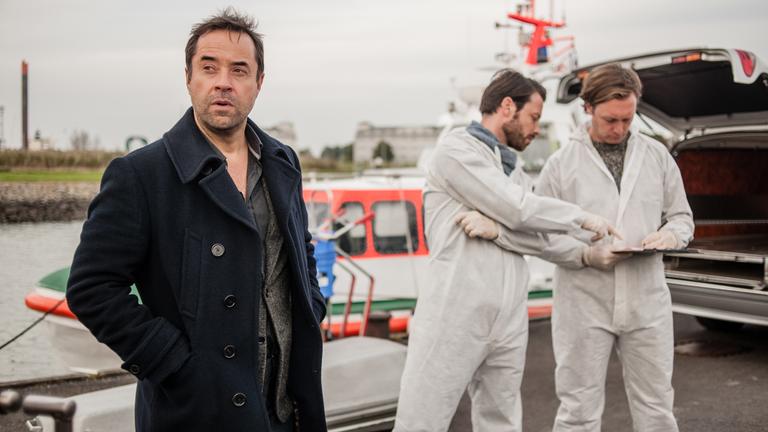 "Mörderische Stille": Hauptkommissar Holzer (Jan Josef Liefers) und zwei Kriminaltechniker (Komparsen) stehen an einer Bootsanlegestelle. Im Hintergrund liegen ein Sarg und ein Polizeischiff.