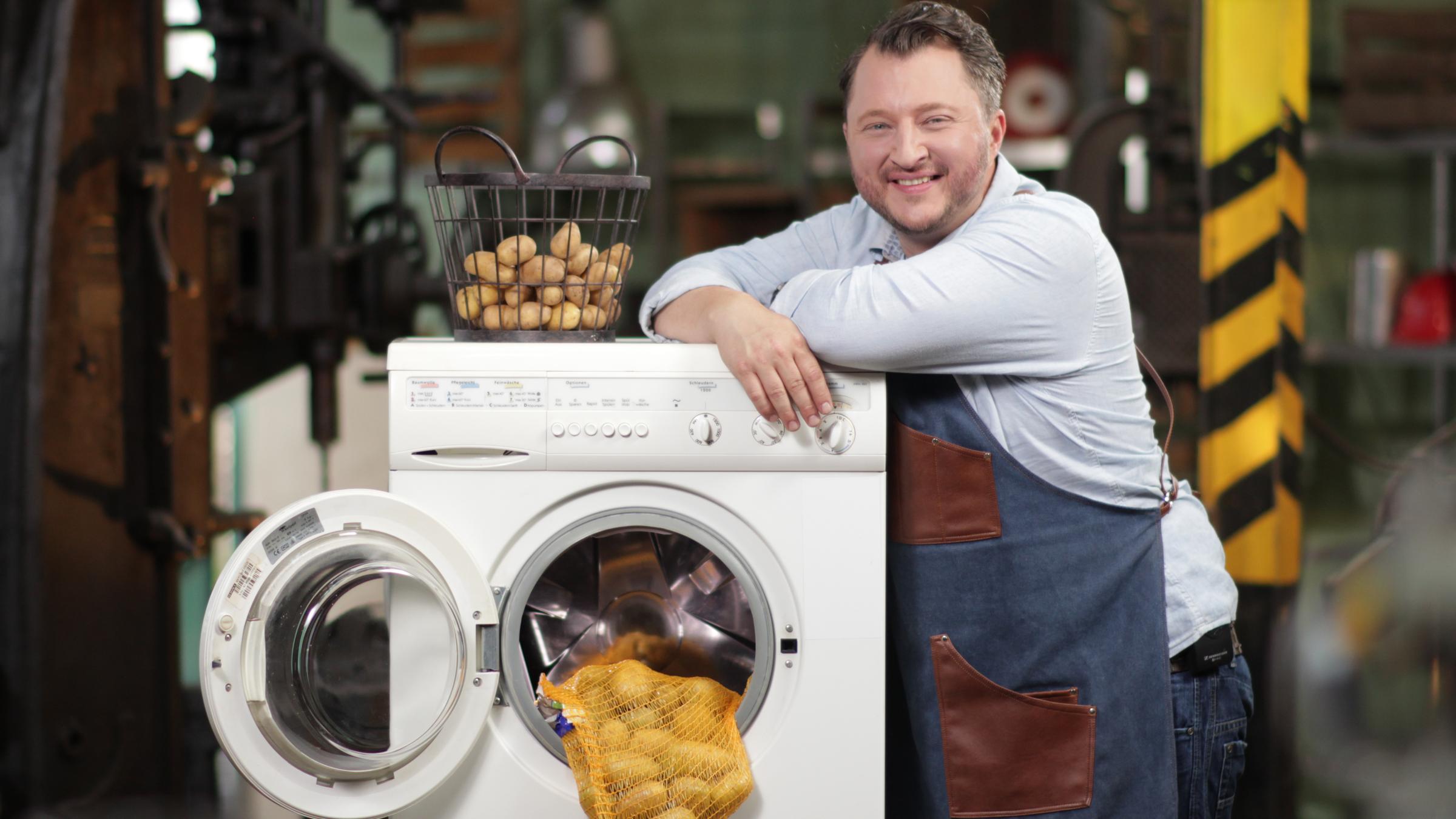 "ZDFzeit: Die Tricks der Lebensmittelindustrie. Günstig und gesund (1)": Sebastian Lege lehnt an einer Waschmaschine, aus der ein Netz mit Kartoffeln hängt.
