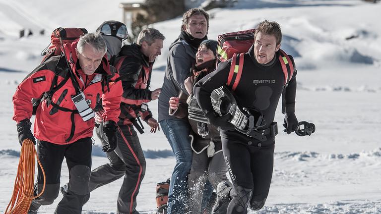 "Der Bergdoktor - Gestohlenes Glück": Zwei Einsatzkräfte der Bergrettung laufen gemeinsam mit Hans Gruber (Heiko Ruprecht) über Eis und Schnee. Hans trägt Taucherflossen unterm Arm. Im Hintergrund hält Dr. Martin Gruber (Hans Sigl) die schreiende Sabine Moser (Janina Hartwig) in den Armen.