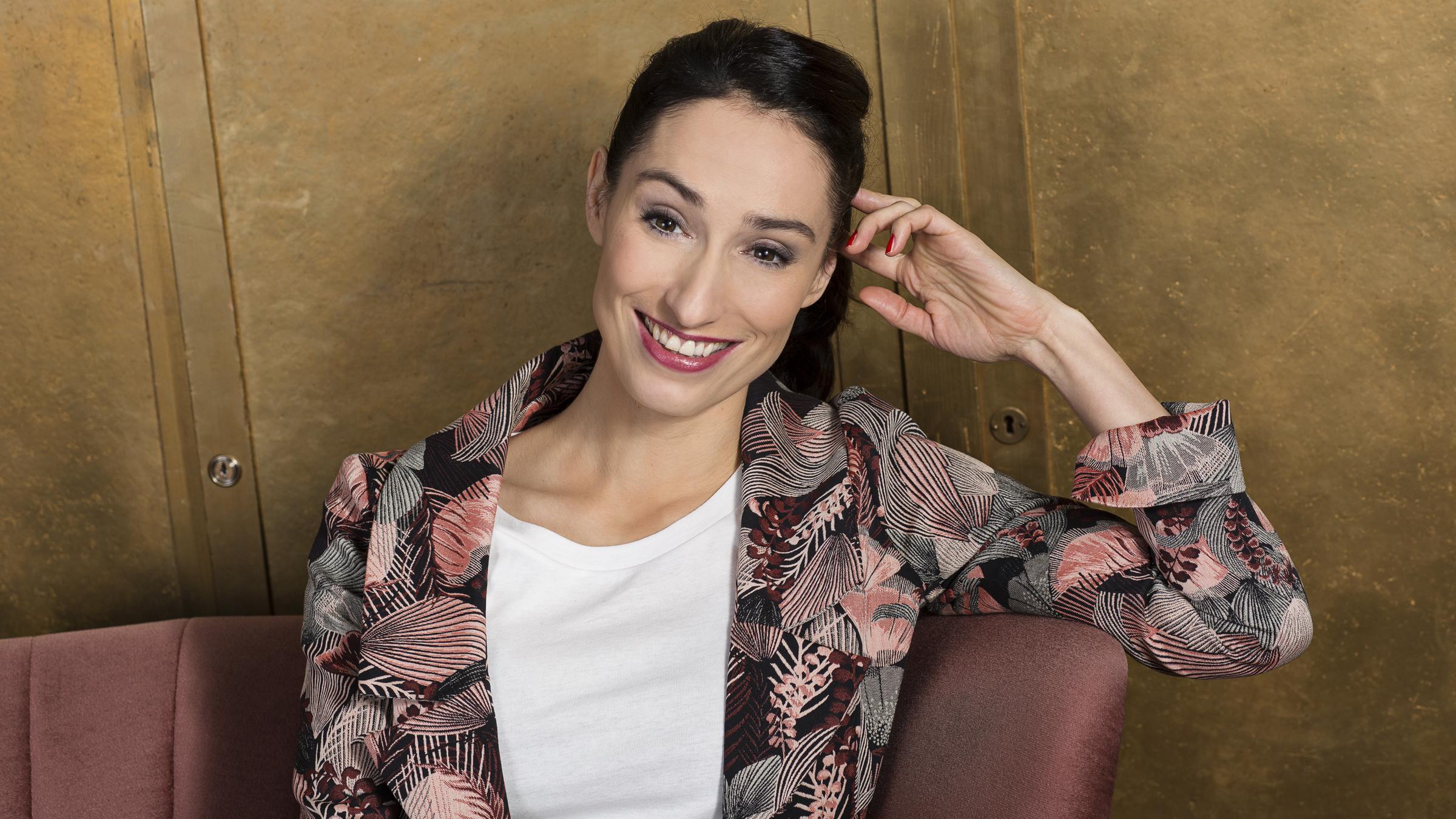 "Kulturzeit - Moderatorin Vivian Perkovic ab 30. Januar 2017": Vivian Perkovic sitzt lächelnd auf einer rosafarbenen Couch. Sie trägt eine gemusterte Jacke und ein weißes Shirt.