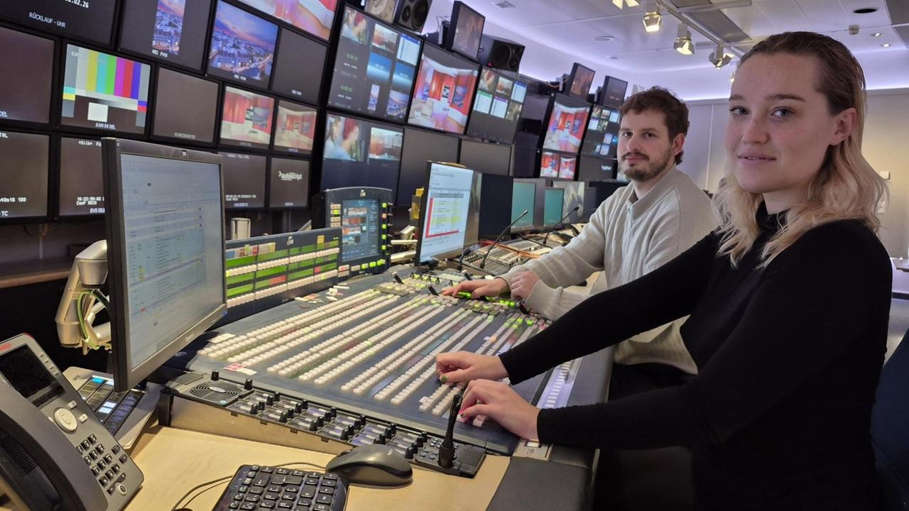 Eine junge Frau und ein junger Mann am Pult in einer Bildregie vor vielen Monitoren lächeln seitlich in die Kamera