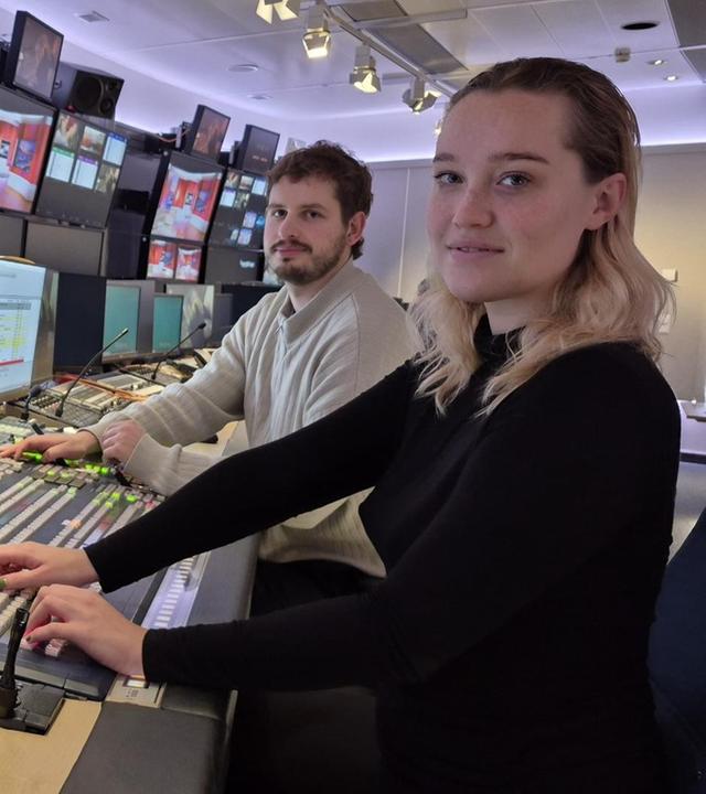 Eine junge Frau und ein junger Mann am Pult in einer Bildregie vor vielen Monitoren lächeln seitlich in die Kamera