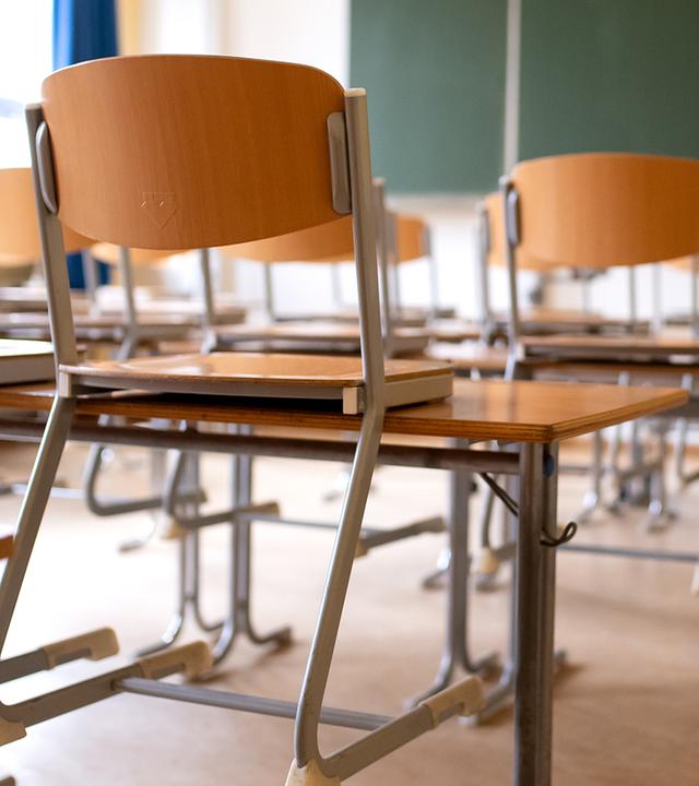 Ein leeres Klassenzimmer. Die Stühle sind auf die Tische gestellt. Im Hintergrund eine grüne Tafel.