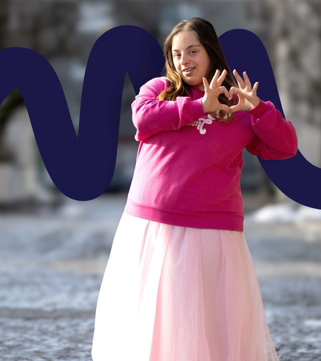 Bilge Asar steht auf einer gepflasterten Straße zwischen Häusern. Sie trägt denselben pinken Pullover mit weißer Schrift und einen rosafarbenen Tüllrock. Mit einer Hand hält sie eine Haarsträhne, die andere stützt sie selbstbewusst in die Hüfte. Der Hintergrund ist unscharf, die Sonne scheint. Sie schaut direkt in die Kamera.