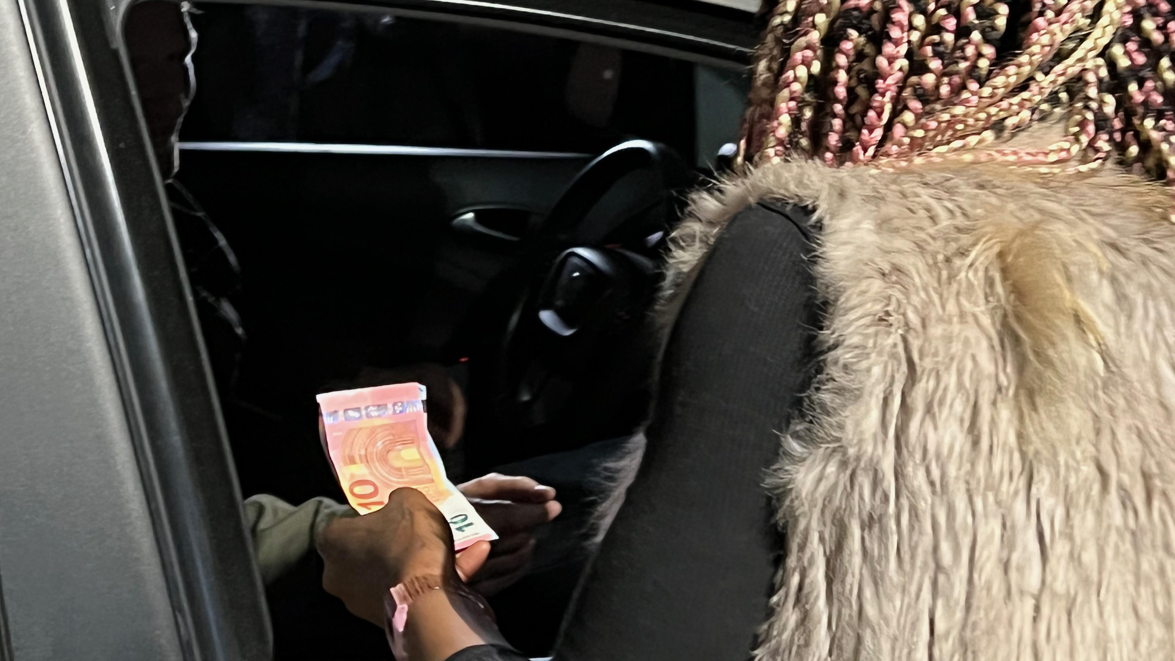 Junge Frau beugt sich auf der Beifahrerseite in ein Auto und erhält Geld von einem kaum sichtbaren Mann im Auto sitzend.
