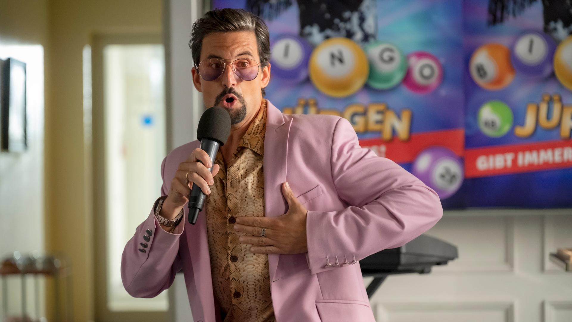 Ein Mann mit goldenem Hemd, rosa Anzug und auffälliger Brille, der in ein Mikrofon spricht.