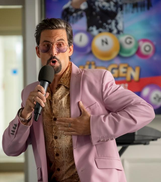 Ein Mann mit goldenem Hemd, rosa Anzug und auffälliger Brille, der in ein Mikrofon spricht.