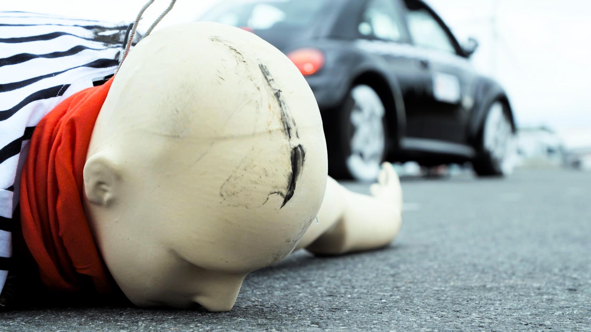 Im Vordergrund liegt ein Biofidel Dummy mit dem Gesicht nach unten auf dem Asphalt (Kopf nah), der Kopf ist oben beschädigt, im Hintergrund (unscharf) steht ein schwarzes Auto.  