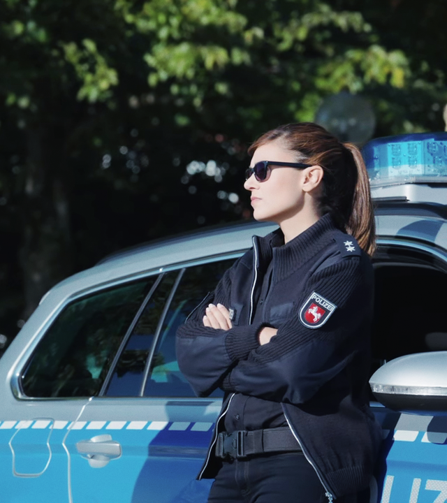 Henk Cassens (Maxim Mehmet) und Süher Özlügül (Sophie Dal) stehen am Polizeiauto, beide in Uniform und Sonnenbrille. 