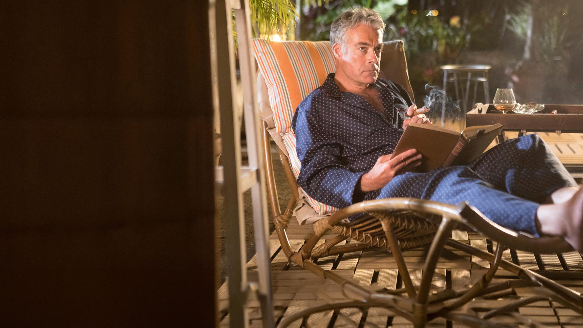"Death in Paradise - Bittere Bohnen": Am Abend. Benedict Dacre (Robert Portal) sitzt im Bademantel auf einer Bambus-Gartenliege auf seiner Terrasse. Er liest ein Buch und raucht eine Zigarre. Auf dem Tisch steht ein Glas Cognac und ein Aschenbecher.
