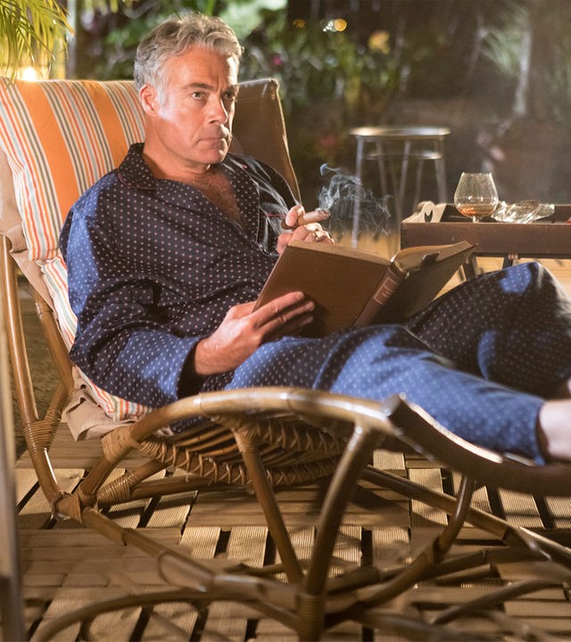 "Death in Paradise - Bittere Bohnen": Am Abend. Benedict Dacre (Robert Portal) sitzt im Bademantel auf einer Bambus-Gartenliege auf seiner Terrasse. Er liest ein Buch und raucht eine Zigarre. Auf dem Tisch steht ein Glas Cognac und ein Aschenbecher.
