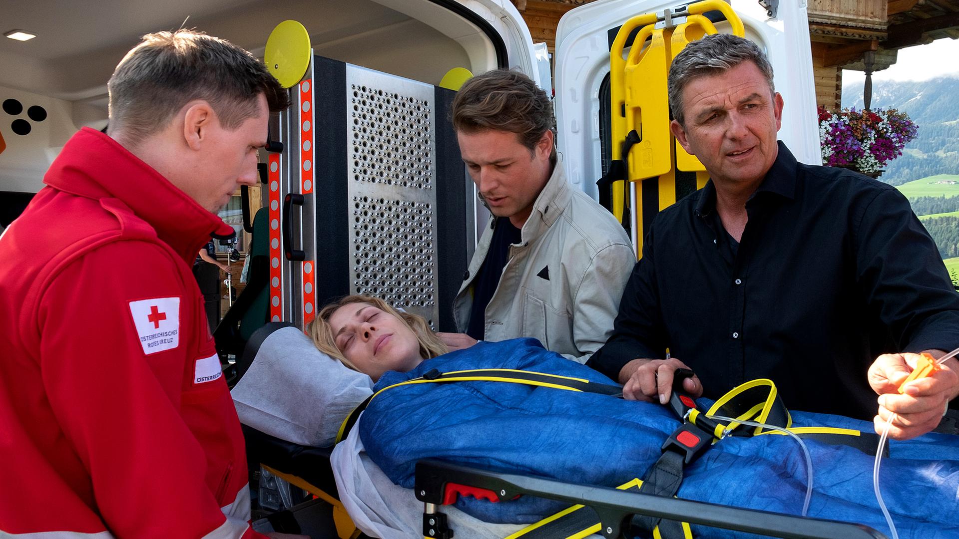 Ein Sanitäter (Komparse), Dr. Martin Gruber (Hans Sigl) und Jan Wöhlert (Julian Looman) heben Lara Wöhlert (Katharina Schlothauer) auf einer Liege in den Krankenwagen.