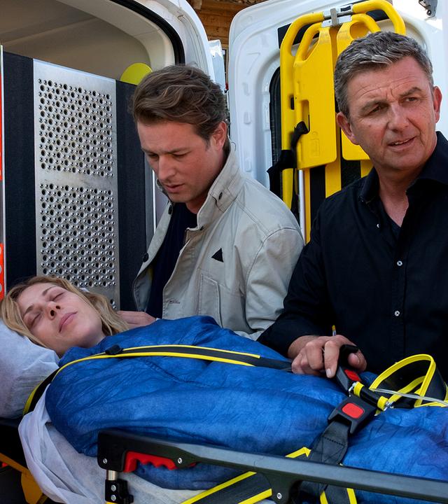 Ein Sanitäter (Komparse), Dr. Martin Gruber (Hans Sigl) und Jan Wöhlert (Julian Looman) heben Lara Wöhlert (Katharina Schlothauer) auf einer Liege in den Krankenwagen.