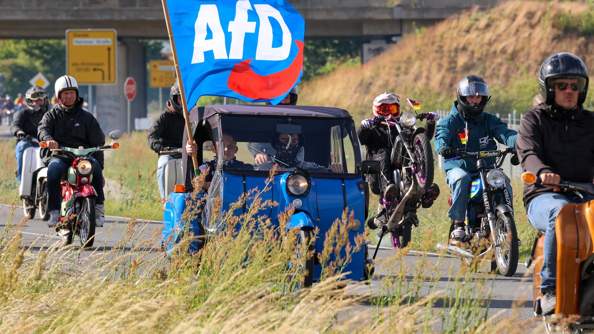 Mopedfahrer fahren gemeinsam mit einem blauen Kleinstwagen, aus dem jemand eine AfD-Flagge aus dem Fenster hält.