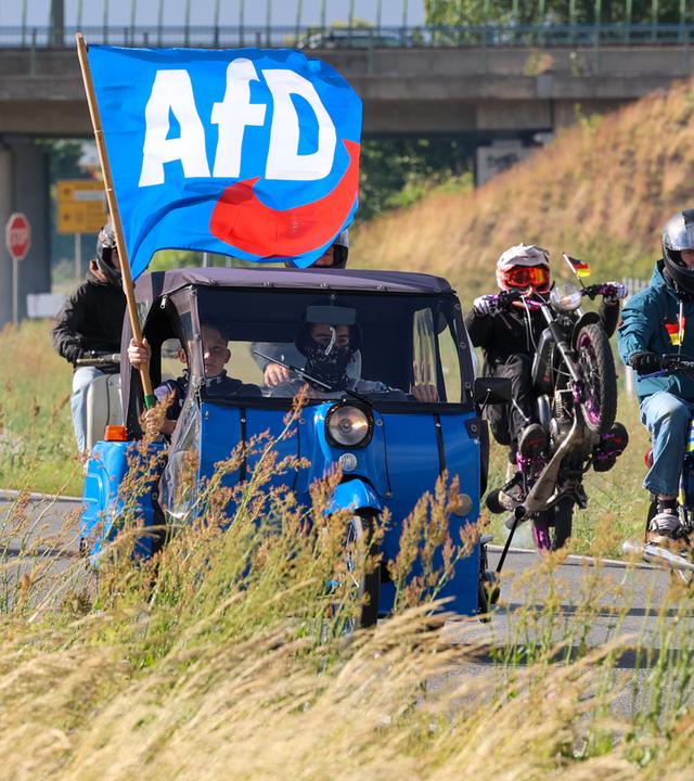 Mopedfahrer fahren gemeinsam mit einem blauen Kleinstwagen, aus dem jemand eine AfD-Flagge aus dem Fenster hält.