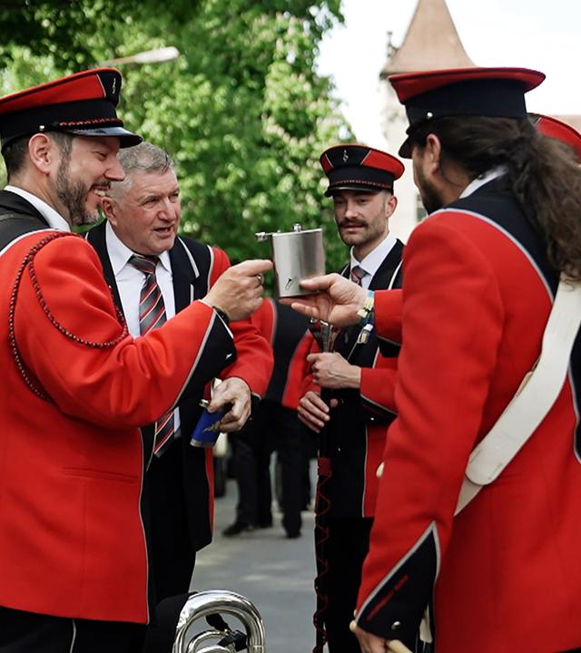 Sechs Männer in rot-schwarzen Jacken mit Musikinstrumenten. Ein Flachmann wird im Zentrum der Gruppe herumgereicht.