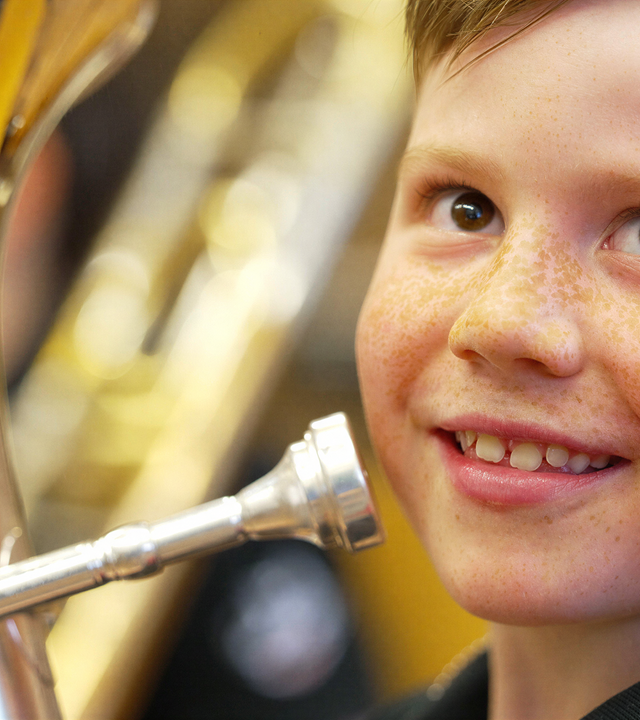 Das Bild zeigt einen Jungen, der ein Musikinstrument, vermutlich eine Tuba, hält. Er hat eine fröhliche Ausstrahlung und lächelt. Der Junge ist möglicherweise zwischen 8 und 12 Jahren alt und hat kurze, blonde Haare. Sein Gesicht ist von Sommersprossen übersät, und er trägt ein schwarzes T-Shirt. Er hält das Mundstück der Tuba in der Nähe seines Gesichts. Im Hintergrund sind unscharfe Silhouetten anderer Musikinstrumente und möglicherweise weiterer Personen zu erkennen, die ebenfalls Musikinstrumente spielen oder vorbereiten. Die Beleuchtung im Raum ist warm und freundlich. Das Bild vermittelt eine Stimmung der Vorfreude und des gemeinschaftlichen Musizierens.