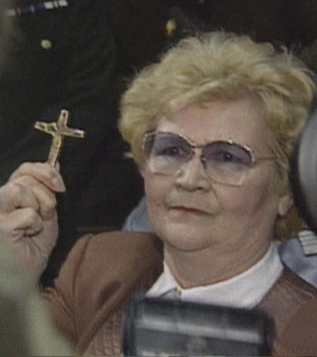 Eine Frau, ca. 65 Jahre alt in einem Gerichtsaal hält in ihrer rechten Hand ein goldenes Kreuz in Richtung zahlreicher Kameraleute. Links daneben: Lichtschematische Darstellung eines Fingerabdrucks.