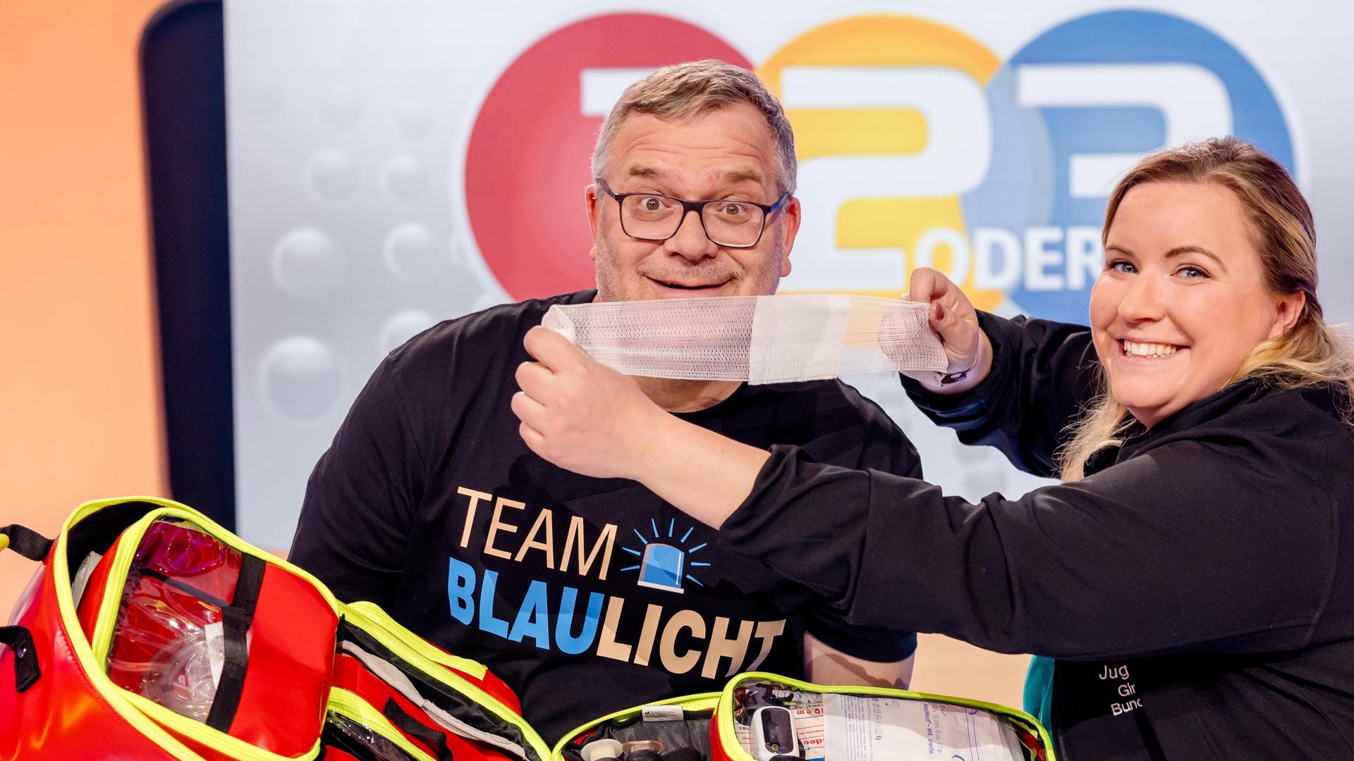 Elton steht mit einer Erste-Hilfe-Tasche im Studio und präsentiert deren Inhalt. Gina steht daneben und tut lachend so, als würde sie Elton einen Verband anlegen.