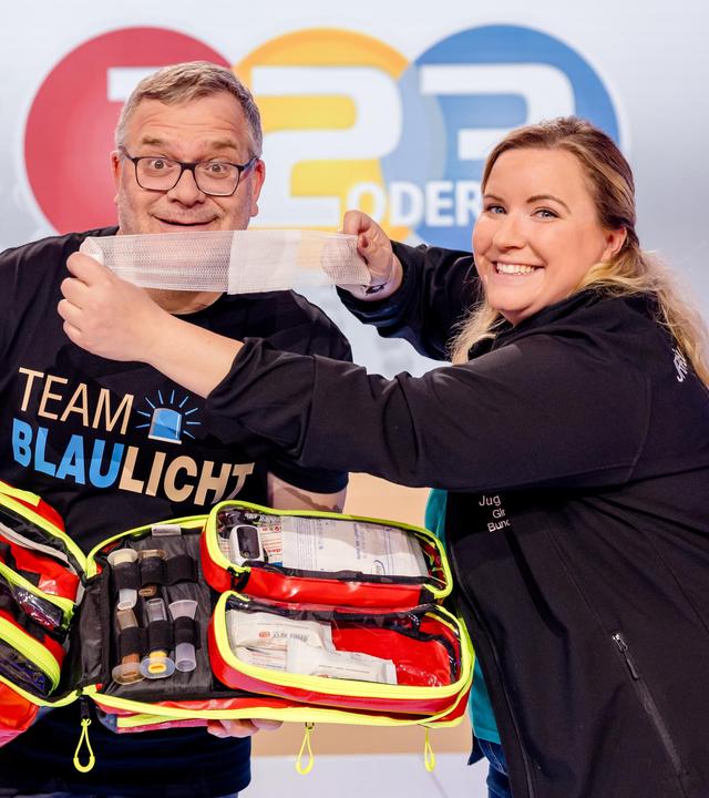Elton steht mit einer Erste-Hilfe-Tasche im Studio und präsentiert deren Inhalt. Gina steht daneben und tut lachend so, als würde sie Elton einen Verband anlegen.