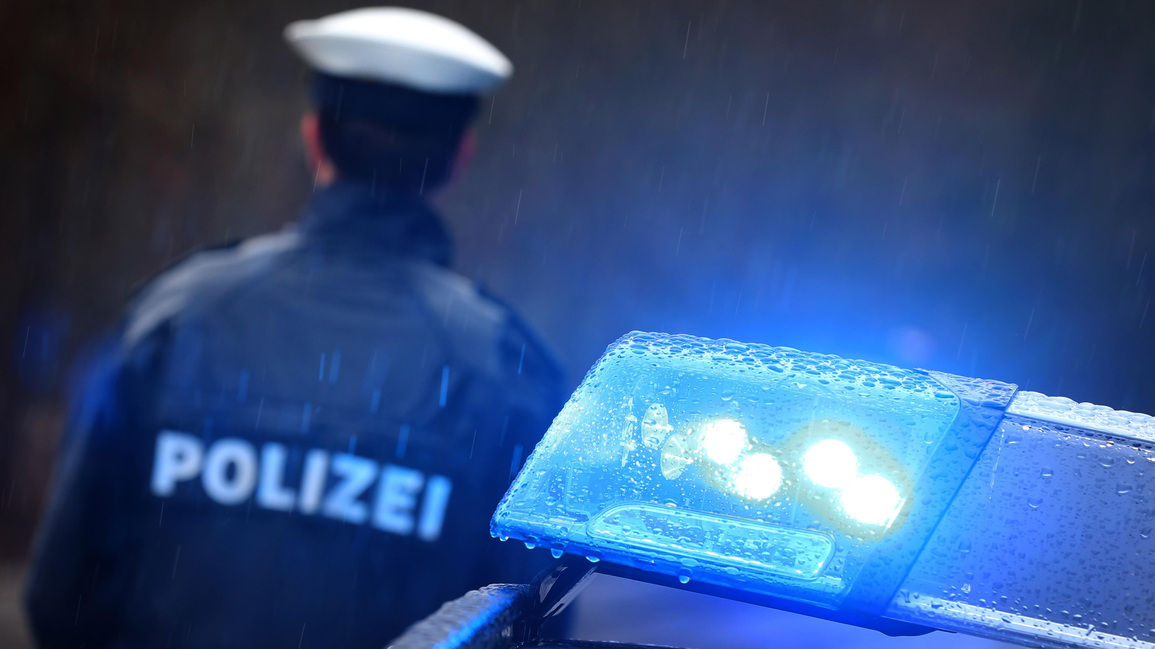 Polizist steht mit dem Rücken zu einem Polizeiauto mit eingeschaltetem Blaulicht 