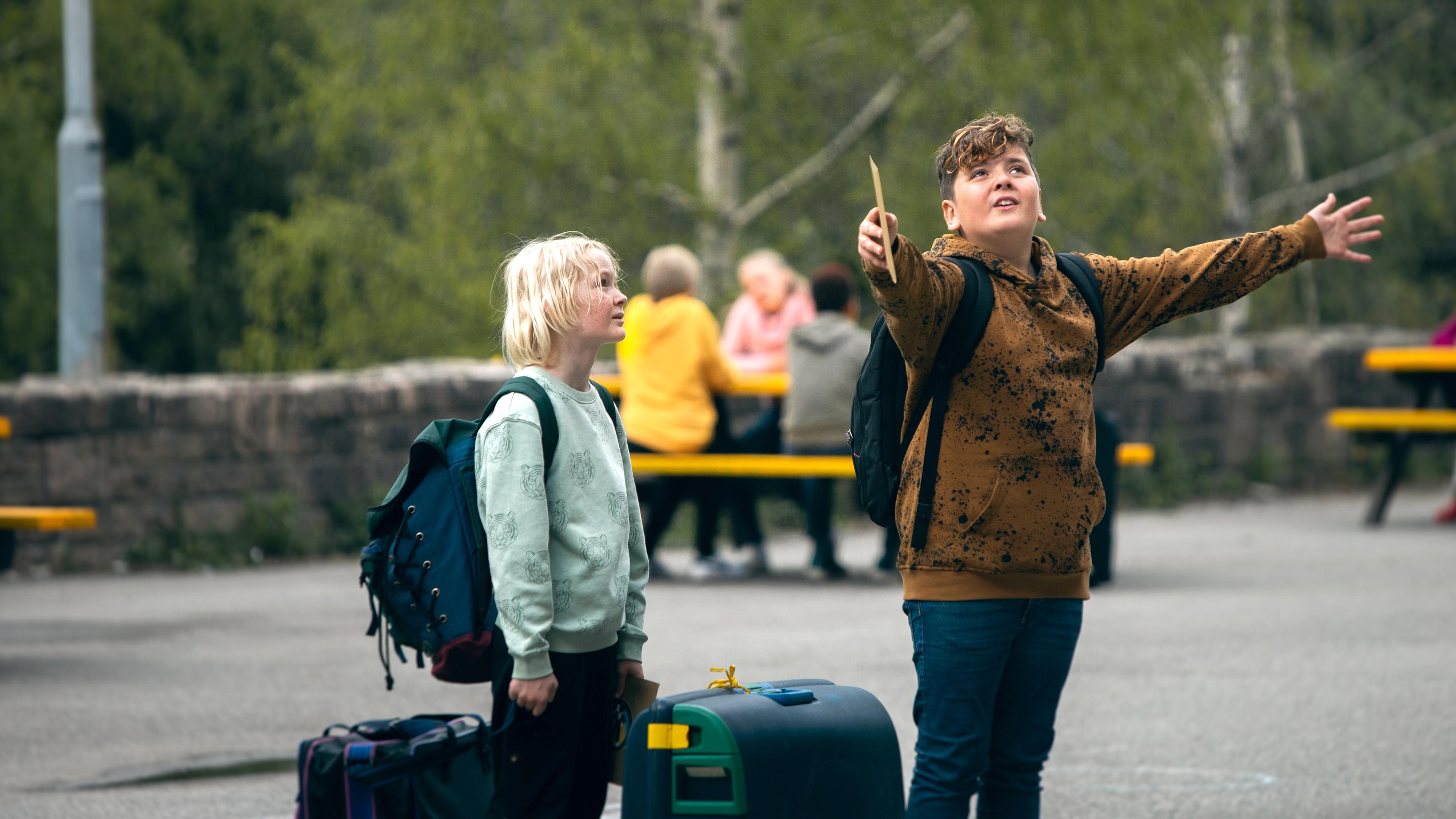 Blaze steht mit ausgestreckten Armen und begeistertem, nach oben gehenden Blick auf dem Schulhof. Links neben ihm steht Raymond, der seine Arme am Körper herunterhängen lässt und skeptisch dreinblickt. Beide tragen einen Rucksack und haben jeweils einen Koffer neben sich stehen.