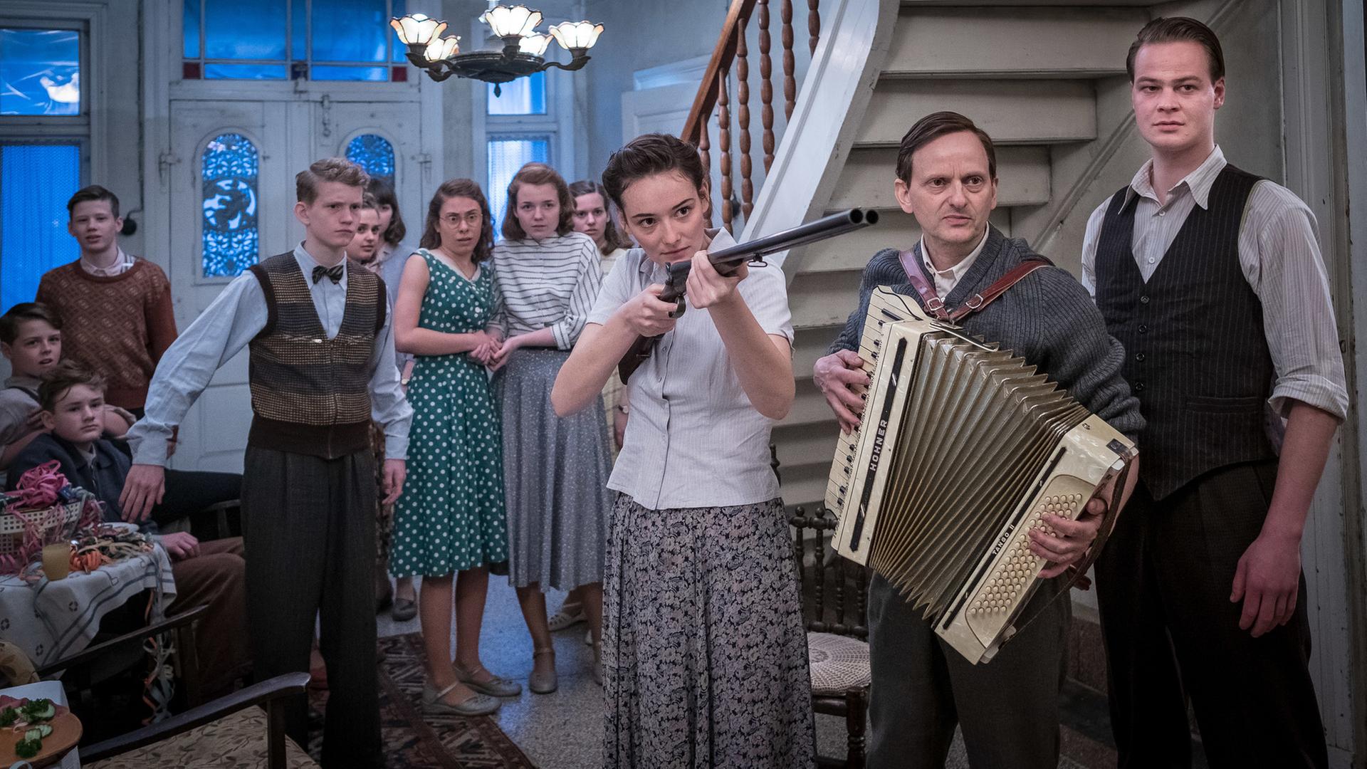 Die junge Vera (Maria Ehrich, Mitte) hält eine Pistole in der Hand. Sie steht in der Eingangshalle eines Gutshauses und ist umgeben von Menschen, der Mann neben ihr (Milan Peschel, rechts) hält ein Akkordeon in den Händen.