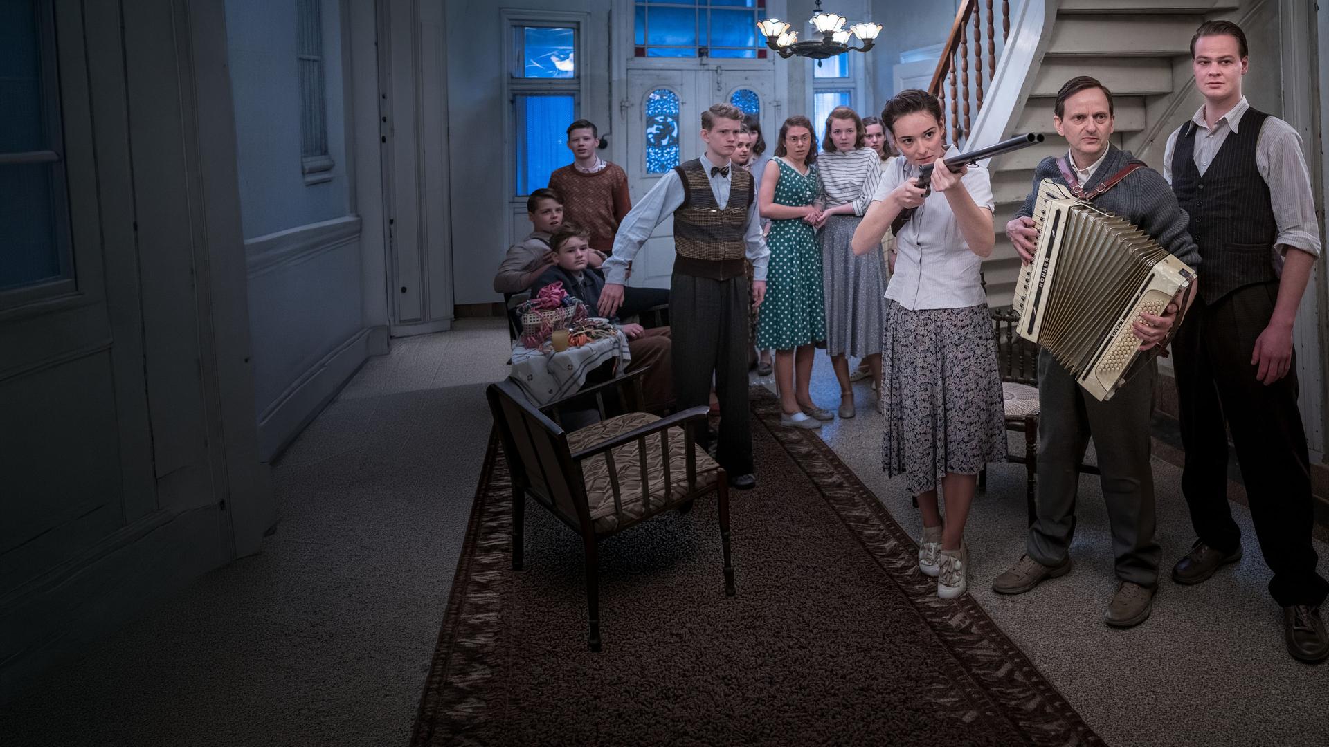 Die junge Vera (Maria Ehrich, Mitte) hält eine Pistole in der Hand. Sie steht in der Eingangshalle eines Gutshauses und ist umgeben von Menschen, der Mann neben ihr (Milan Peschel, rechts) hält ein Akkordeon in den Händen.