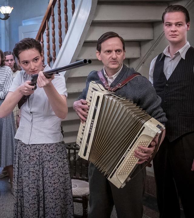 Die junge Vera (Maria Ehrich, Mitte) hält eine Pistole in der Hand. Sie steht in der Eingangshalle eines Gutshauses und ist umgeben von Menschen, der Mann neben ihr (Milan Peschel, rechts) hält ein Akkordeon in den Händen.