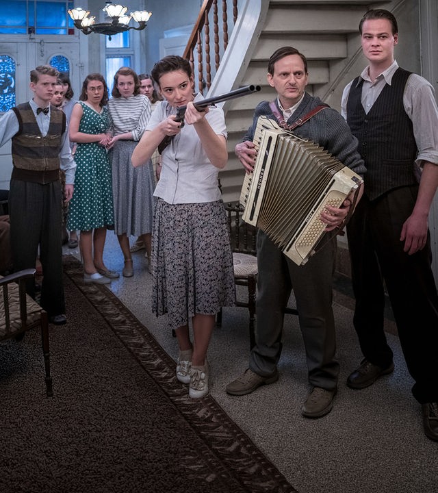 Die junge Vera (Maria Ehrich, Mitte) hält eine Pistole in der Hand. Sie steht in der Eingangshalle eines Gutshauses und ist umgeben von Menschen, der Mann neben ihr (Milan Peschel, rechts) hält ein Akkordeon in den Händen.