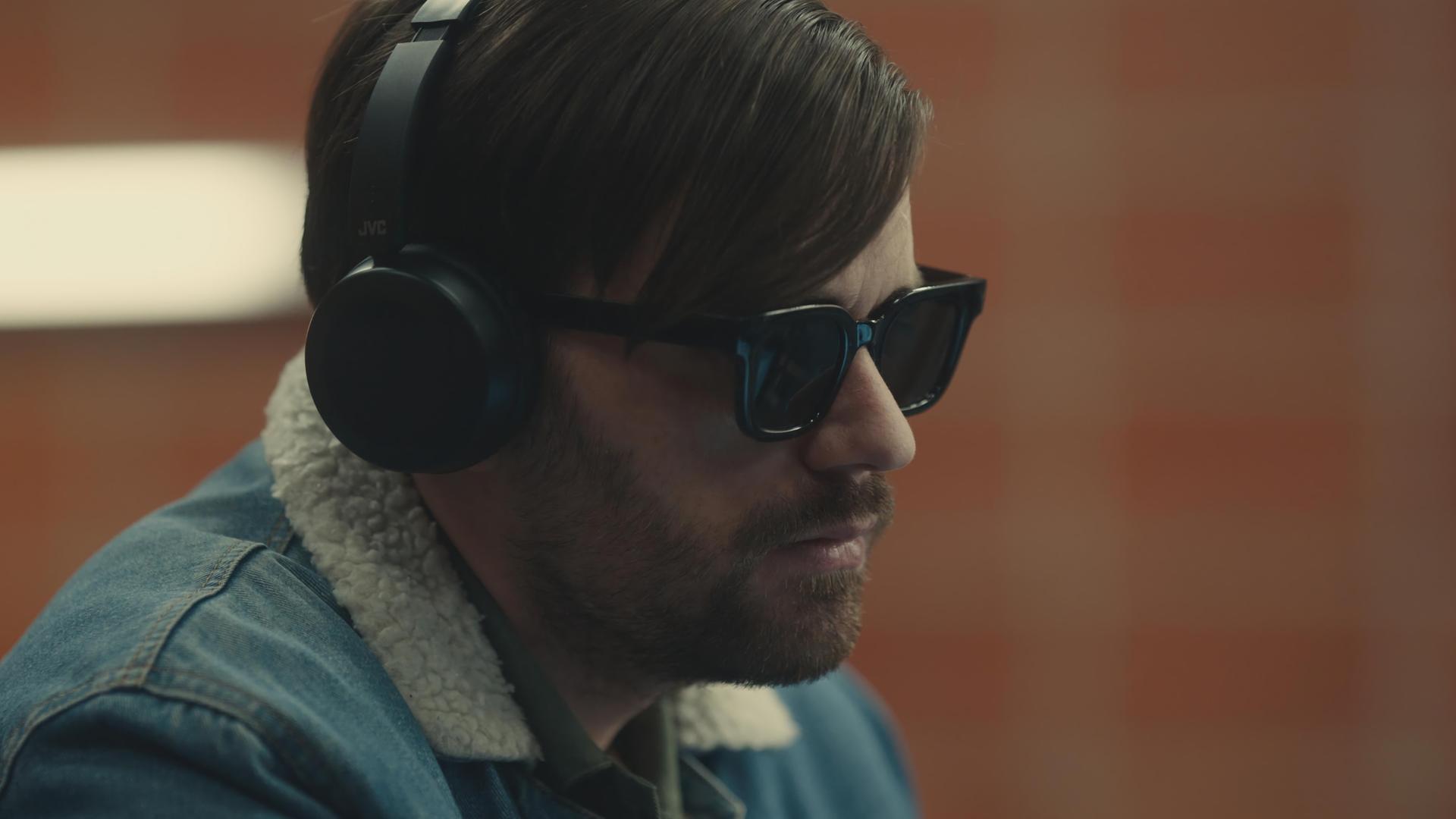 "Blind Sherlock - Ich höre was, was du nicht hörst": Roman (Bart Kelchtermans) sitzt in einem Raum. Er trägt eine dunkle Brille und Kopfhörer und schaut zur Seite.
