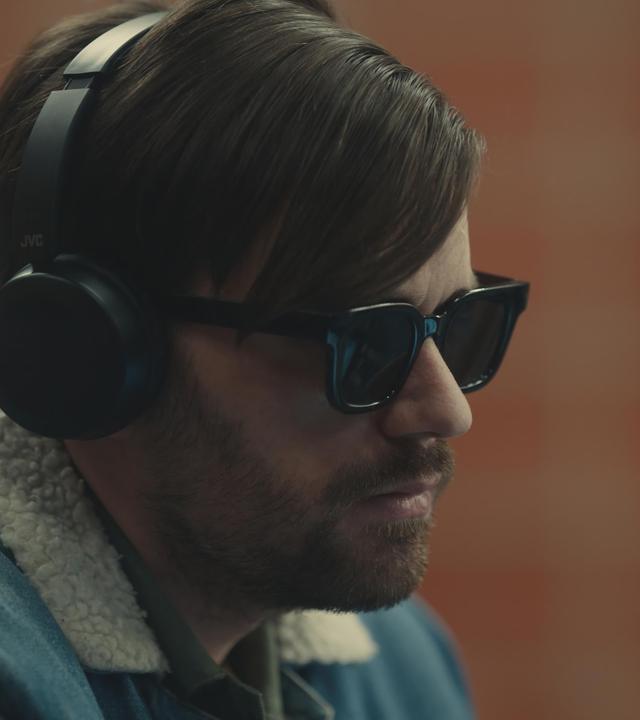 "Blind Sherlock - Ich höre was, was du nicht hörst": Roman (Bart Kelchtermans) sitzt in einem Raum. Er trägt eine dunkle Brille und Kopfhörer und schaut zur Seite.