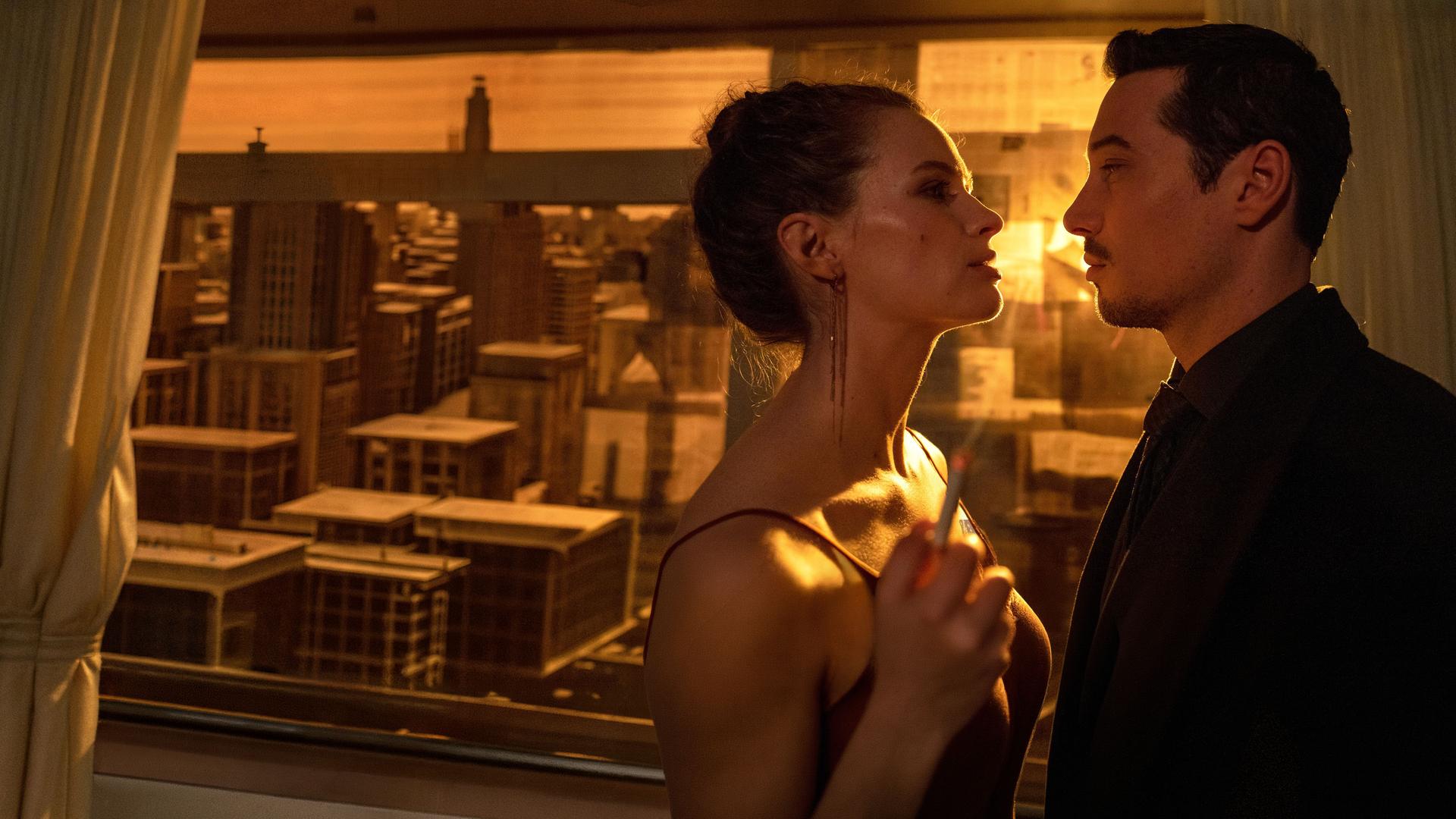 "Blindspot": Anara van Veeren (Marlene Tanczik) und Max van Veeren (Klaus Steinbacher) stehen sich eng gegenüber und schauen sich gegenseitig mit ernster Miene an. Anara hält in ihrer rechten Hand eine brennende Zigarette. Durch das mit Zeitungspapier verdunkelte Fenster scheint gelbes Licht.