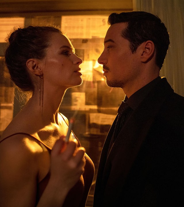 "Blindspot": Anara van Veeren (Marlene Tanczik) und Max van Veeren (Klaus Steinbacher) stehen sich eng gegenüber und schauen sich gegenseitig mit ernster Miene an. Anara hält in ihrer rechten Hand eine brennende Zigarette. Durch das mit Zeitungspapier verdunkelte Fenster scheint gelbes Licht.