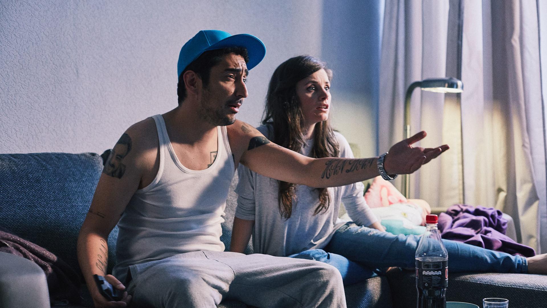 Jessica und Sol sitzen auf der Couch. Sol zeigt aufgebracht in Richtung Fernseher.