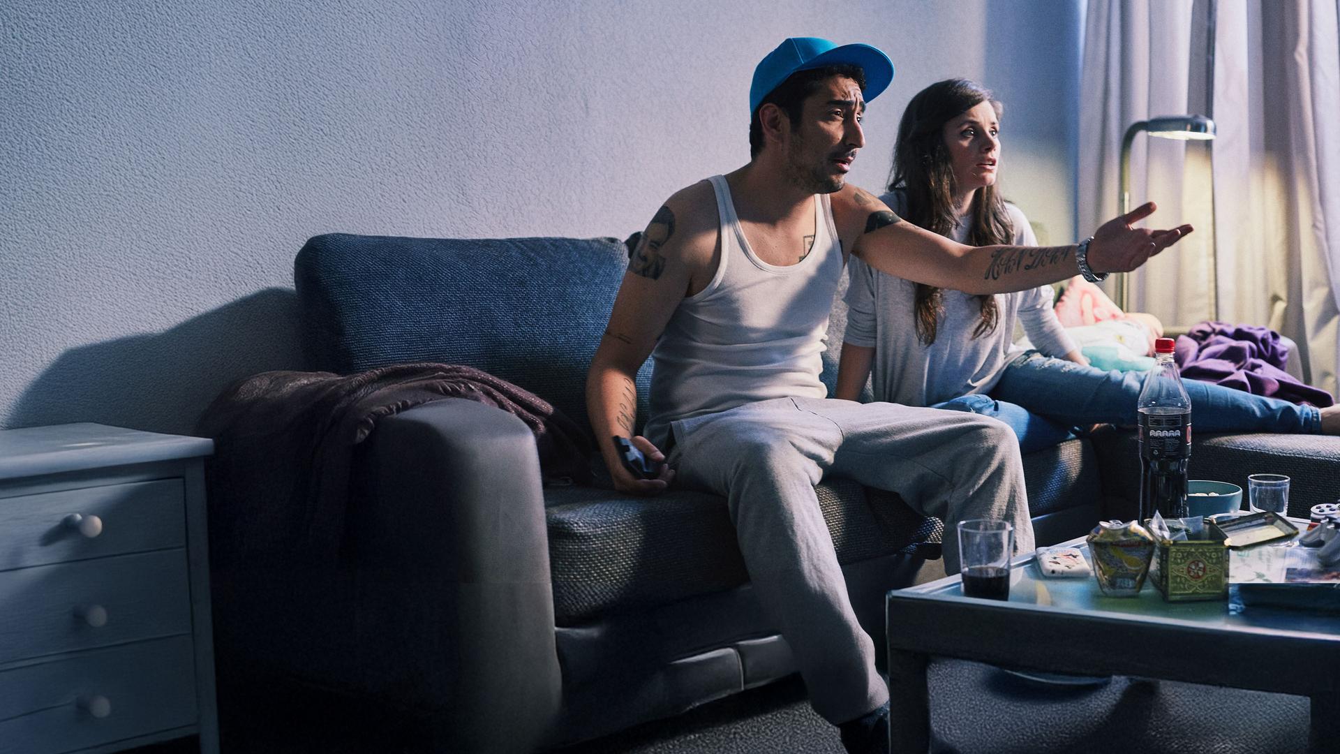 Jessica und Sol sitzen auf der Couch. Sol zeigt aufgebracht in Richtung Fernseher.