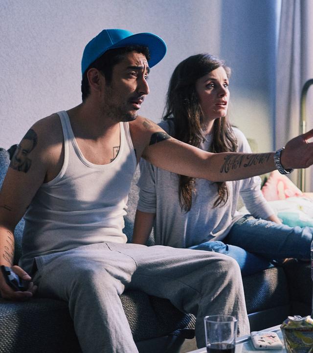 Jessica und Sol sitzen auf der Couch. Sol zeigt aufgebracht in Richtung Fernseher.