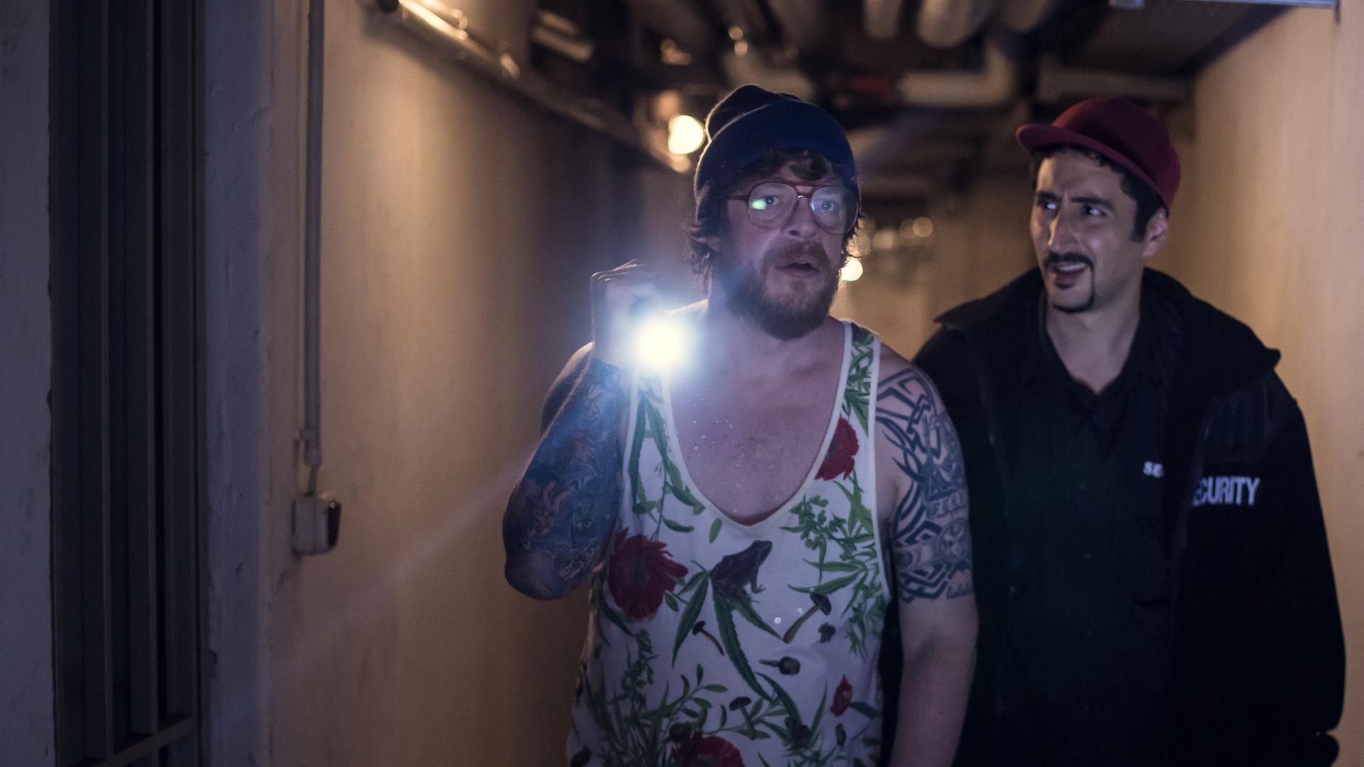 Hardy, mit Taschenlampe, und Sol in Securtity-Jacke gehen einen dunklen Gang entlang.