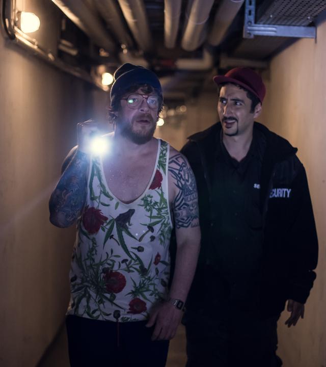 Hardy, mit Taschenlampe, und Sol in Securtity-Jacke gehen einen dunklen Gang entlang.