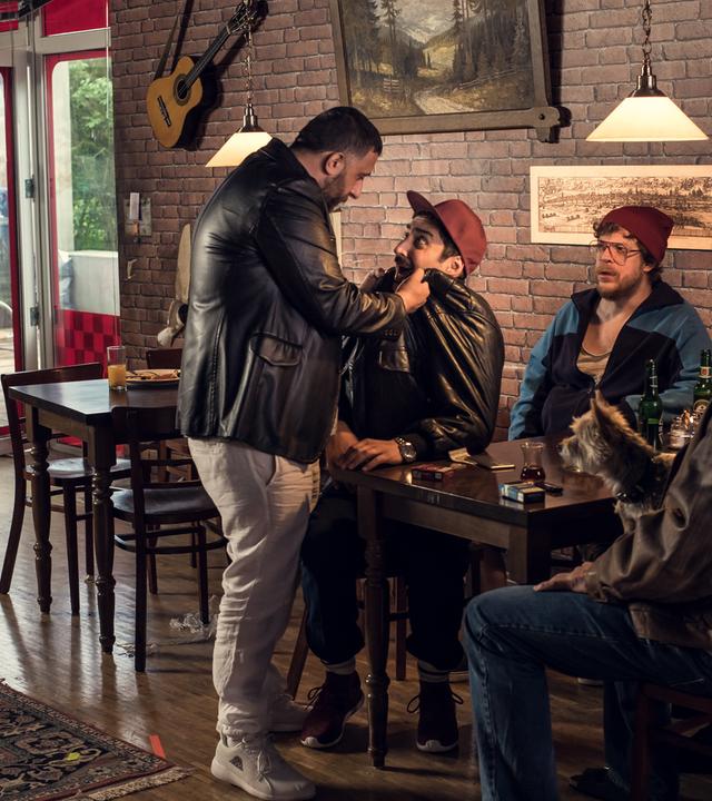 Hardy und Sol sitzen in einer schummerig ausgeläuchteten Bar an einem Tisch. Vor Sol steht Volkan und greift ihn bedrohlich an der Lederjacke.