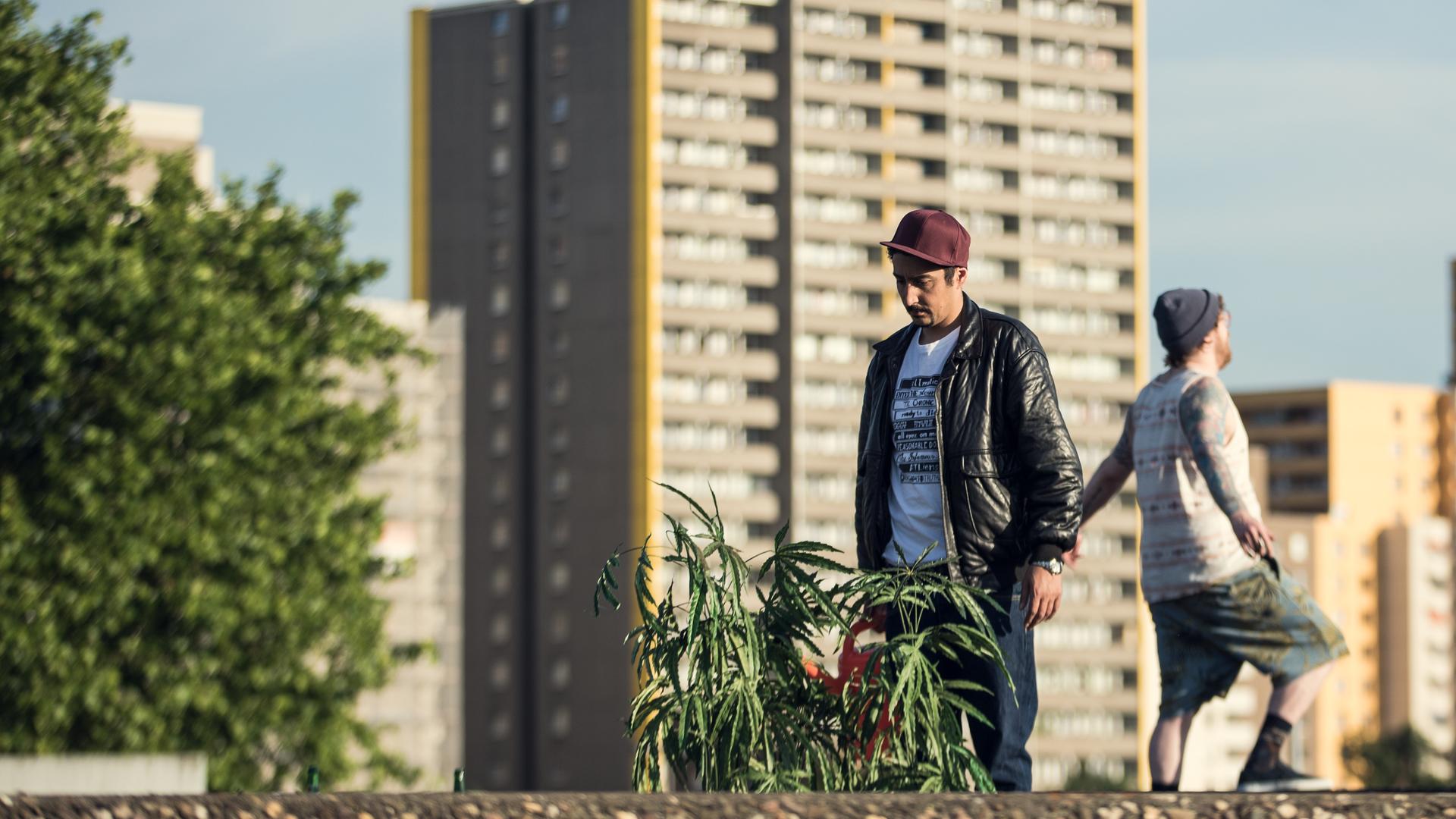 Sol und Hardy haben sich den Rücken zugewandt und stehen im Abstand von einem Meter zueinander. Im Hintergrund ist eine Hochhausreihe Kölns zu sehen.
