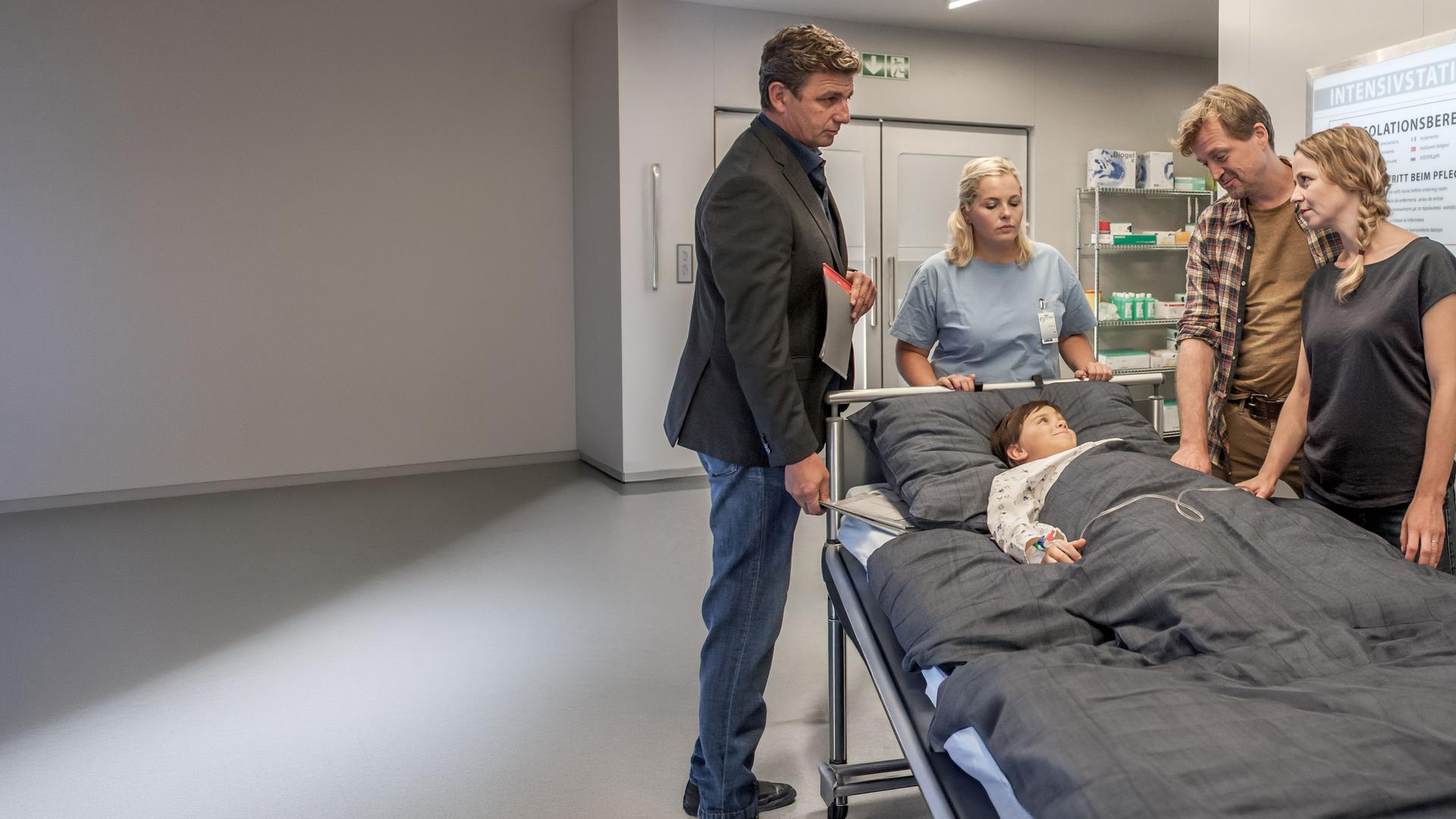 "Der Bergdoktor - Blut und Wasser (2)": Dr. Martin Gruber (Hans Sigl), Daniel (Patrick von Blume) und Petra Förster (Theresa Scholze) stehen im Krankenhausflur um das Bett von Finn (Arian Montgomery).