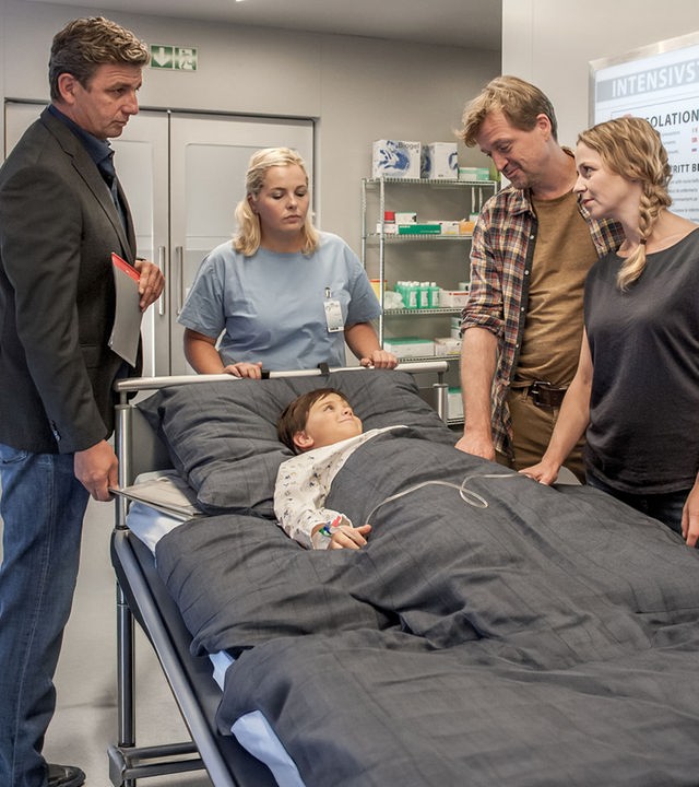 "Der Bergdoktor - Blut und Wasser (2)": Dr. Martin Gruber (Hans Sigl), Daniel (Patrick von Blume) und Petra Förster (Theresa Scholze) stehen im Krankenhausflur um das Bett von Finn (Arian Montgomery).