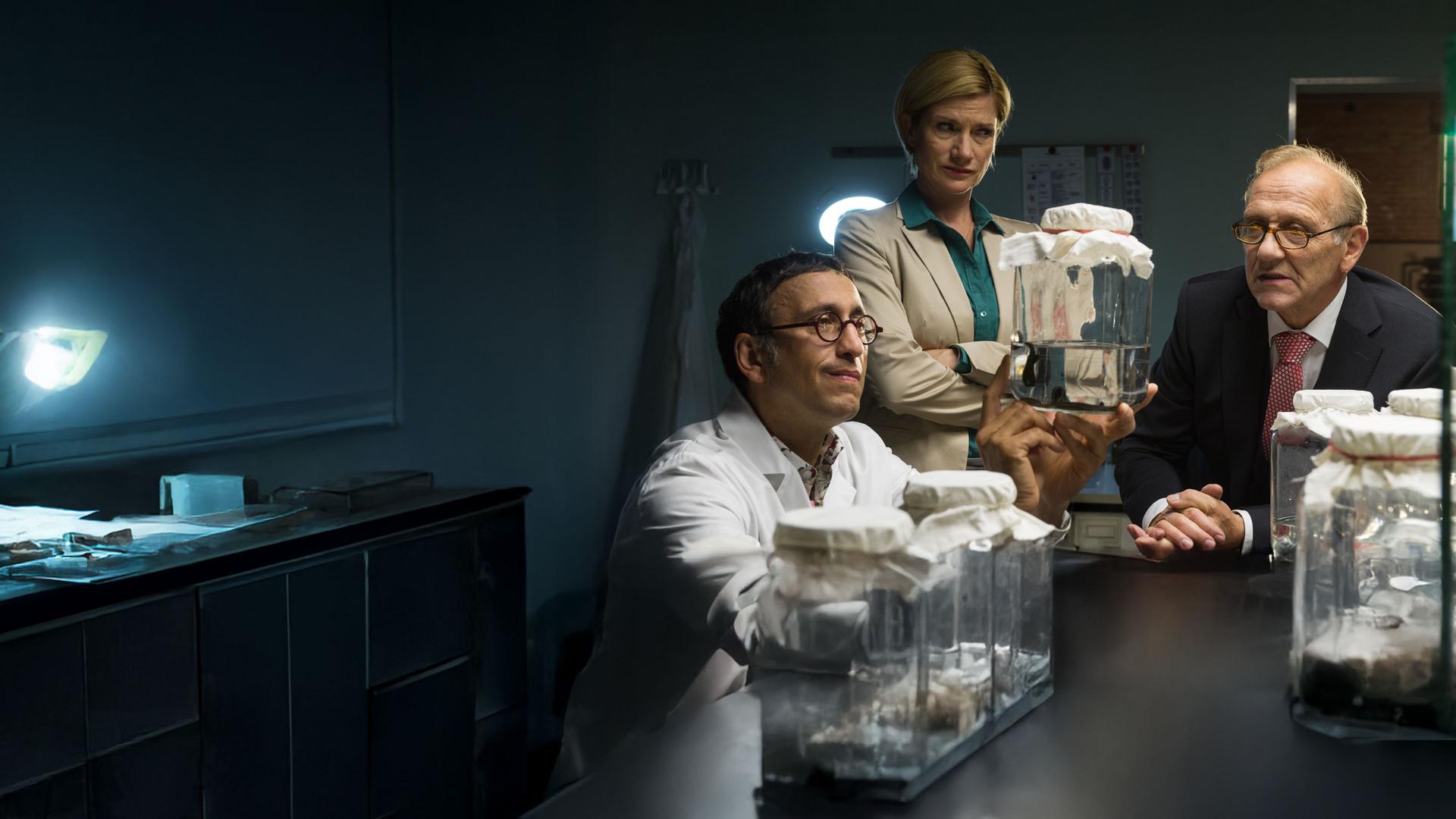 "SOKO Stuttgart - Blutsauger": Martina Seiffert (Astrid M. Fünderich) und Michael Kaiser (Karl Kranzkowski) sitzen in Jan Arnauds (Mike Zaka Sommerfeldt) Labor. Jan hält ein großes abgedecktes Glas in die Höhe darin befinden sich Wasser und Blutegel.